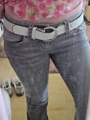 Vitt bälte med glittrande spänne - Snyggt vitt bälte med ett runt spänne dekorerat med glittrande stenar. Bältet har svarta detaljer längs kanterna och är tillverkat i skinnimitation. Jag fick ärva detta bälte från min mamma så jag vet tyvärr inte märket. Bältet är mycket använt men i helt okej skick. 