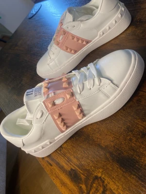 Valentino vita sneakers med rosa nitar - Snygga vita sneakers från Valentino med breda rosa band över vristen dekorerade med ikoniska pyramidformade nitar. Skorna har en chunky sula och är tillverkade i slätt läder med klassisk låg silhuett. Perfekta för dig som vill sticka ut med en lyxig touch.