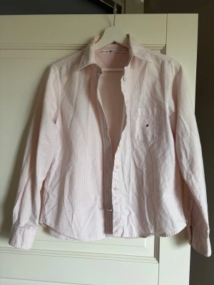 Rosa och vit randig skjorta Tommy Hilfiger - Klassisk skjorta från Tommy Hilfiger i ljusrosa och vitt med tunna ränder. Skjortan har lång ärm, bröstficka med liten logga och knappar framtill. Tillverkad i bomull och har en normal passform. Perfekt för en clean och stilren look.