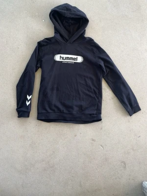 Svart hoodie från Hummel - Snygg svart hoodie från Hummel med vit logga på bröstet och klassiska vita pilar på ena ärmen. Tillverkad i mjukt material och har en skön huva. Perfekt för en sportig och avslappnad stil.