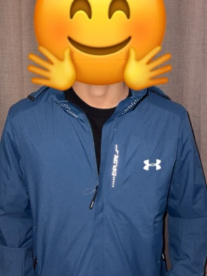 Blå vindjacka från Under Armour - Snygg blå vindjacka från Under Armour med dragkedja och huva. Jackan har vit logga på bröstet och justerbar huva. Perfekt för blåsiga dagar och enkel att matcha med andra plagg.