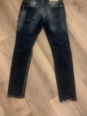 Mörkblå skinny jeans från Dsquared2 - Säljer ett par mörkblå skinny jeans från Dsquared2 med snygga slitningar och klassiska bakfickor. Jeansen har en cool patch med Dsquared2-logga och D8Q2 i grönt. Perfekta för dig som gillar streetstyle och trendiga detaljer.