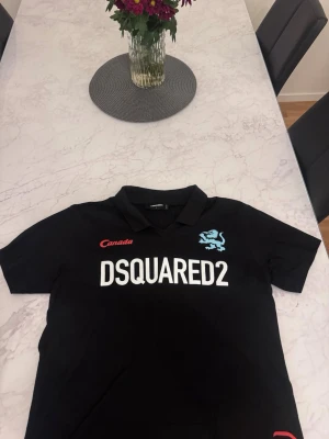 Svart DSQUARED2 piké med tryck - Svart pikétröja från DSQUARED2 med vit logga framtill, röd 'Canada'-text och blått lejontryck. På ryggen står det 'CATEN' och en stor blå siffra 2. Klassisk krage och korta ärmar. Perfekt för dig som gillar streetwear och tydliga loggor.