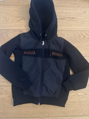 Svart hoodie från Hugo Boss - En snygg svart hoodie av hugo boss med snygg orange text och har även byxorna till den (blir dyrare) säljer den för jag råka ta en liten storlek!