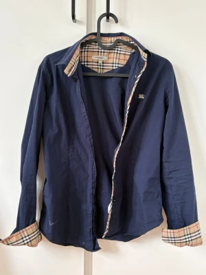 Blå skjorta från Burberry med rutiga detaljer - Snygg mörkblå skjorta från Burberry med klassiska rutiga detaljer på insidan av krage och ärmslut. Skjortan har knappar framtill och ett broderat Burberry-emblem på bröstet. Perfekt för dig som gillar stilrena och exklusiva plagg.OBS storleken är väldigt liten passar mig som är 166.