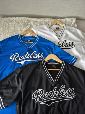 Sommar jerseys  - Säljer en blå baseball-inspirerad t-shirt från FSBN med texten 'Reckless' broderad framtill. T-shirten har korta ärmar, knappar framtill och vita detaljer på krage och ärmslut. Perfekt för dig som vill sticka ut med sportig stil, man får alla 3 på köpet. 
