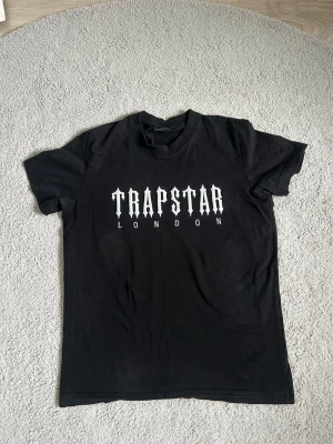 Svart Trapstar London t-shirt - Säljer en stilren svart t-shirt från Trapstar London. T-shirten är i utmärkt skick och perfekt för en snygg look. Den är i storlek M men kan också passa storlek S. Jag är aktiv 24/7 och svarar gärna på alla frågor. Frakten kan göras samma dag eller nästa dag, så du får din t-shirt snabbt! Missa inte chansen att köpa! Tveka inte att höra av dig!