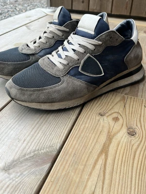 Gråblå sneakers från Philippe Model - Snygga sneakers från Philippe Model i en mix av grå mocka och blå textil. Skorna har vita skosnören, en ikonisk sköld på sidan och en klassisk låg siluett. Yttersulan är svart och vit med bra grepp. Perfekta för en stilren och avslappnad look. Hör av dig vid frågor!