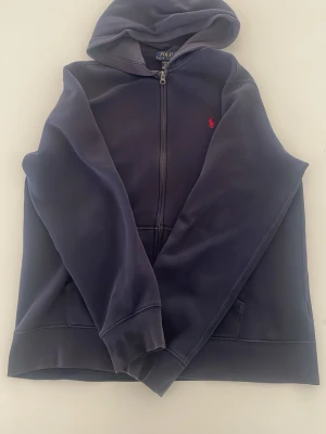 Marinblå hoodie från Polo Ralph Lauren - Klassisk marinblå hoodie från Polo Ralph Lauren med dragkedja framtill och röd logga på bröstet. Tröjan har huva, fickor och långa ärmar. Perfekt för en avslappnad och stilren look.