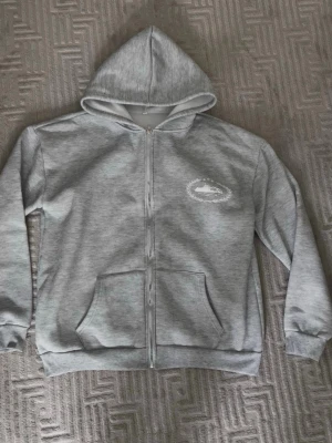Grå zip-hoodie från Corteiz - Snygg grå hoodie med dragkedja från Corteiz. Stor logga i vitt på ryggen och mindre tryck på bröstet. Mjuk bomullskänsla, huva och två fickor framtill. Perfekt för en chill och stilren look.