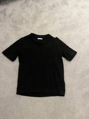 Svart ribbad polotopp från Zara - Säljer en svart ribbad polotopp från Zara i storlek M. Toppen har korta ärmar, klassisk krage och är tillverkad i ett mjukt material som ger en skön passform. Perfekt för en clean och stilren look.