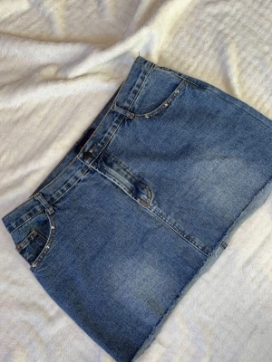 Blå jeanskjol med nitar - Snygg blå jeanskjol i kort modell med rå kant nertill. Kjolen har klassiska fickor fram och bak samt nitar längs fickkanterna. Midjemått: ca 42 cm rakt över