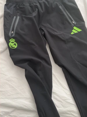Svarta Real Madrid träningsbyxor Adidas - Svarta träningsbyxor från Adidas med Real Madrid-logga i neonfärg och tre ränder på låret. Byxorna har dragkedjor vid fickor och benslut samt elastisk midja. Perfekta för fotbollsträning eller gymmet.