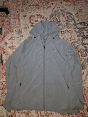 Grå Nike Unlimited Vindjacka - Snygg grå vindjacka från Nike med dragkedja hela vägen, justerbar huva och två sidofickor med dragkedja. Jackan har en minimalistisk design med Nike-logga broderad på bröstet. 2 skador en bredvid loggan en på baksidan, passar m också
