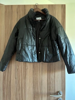 Svart puffer skinnjacka från NA-KD - Säljer en svart puffer skinnjacka från NA-KD med hög krage och tryckknappar framtill. Jackan är kortare i modellen och har två sidofickor. Tillverkad i glansigt konstläder som ger en trendig look.