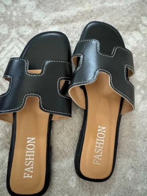 Svarta slip-in sandaler med vita sömmar - Stilrena svarta slip-in sandaler från Fashion med breda remmar i konstläder och vita dekorativa sömmar. Sandalerna har fyrkantig tå och öppen häl, samt en bekväm innersula i beige. Perfekta för en trendig och avslappnad look.