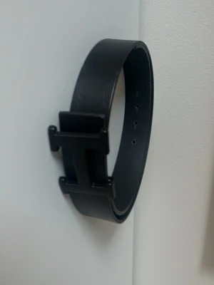 Svart skärp med H-spänne - Snyggt svart bälte med ett stort H-format spänne i matchande svart. Bältet är stilrent och passar perfekt till jeans eller kostymbyxor. Tillverkat i skinn för en klassisk och tidlös look. Perfekt accessoar för att lyfta din outfit.