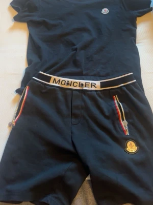 Svart Moncler bodyset med logga - Snyggt svart bodyset från Moncler med matchande t-shirt och shorts. Båda delarna har Moncler-logga, och shortsen har vita och röda detaljer vid dragkedjorna samt ett elastiskt midjeband med Moncler-tryck. Perfekt för en trendig och sportig look.