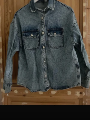 Oversized blå jeansjacka - Snygg oversized jeansjacka i tvättad blå denim med två stora bröstfickor och knappar framtill. Klassisk krage och långa ärmar med knapp vid ärmslut. Perfekt för en avslappnad streetstyle-look.