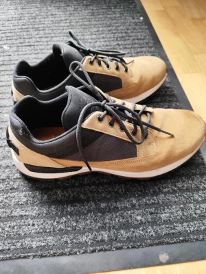 Bruna och svarta sneakers med mockadetaljer - Snygga sneakers i brunt och svart med ovandel i mocka och textil. Skorna har svarta snören, vita sulor och en sportig design. Perfekta för dig som gillar en stilren och avslappnad look.