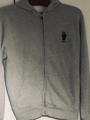 Grå hoodie från Polo Ralph Lauren - Säljer en grå hoodie från Polo Ralph Lauren med dragkedja och klassisk björn-logo på bröstet. Hoodien har långa ärmar, huva och två fickor framtill. Perfekt för dig som gillar stilren och enkel design.
