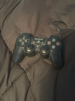 Sony PlayStation DualShock handkontroll - Klassisk svart DualShock handkontroll till PlayStation med kabel. Kontrollen har två analoga spakar, styrkors, fyra färgade knappar och axelknappar. Ergonomisk form i hårdplast, perfekt för gaming på äldre PlayStation-konsoler.