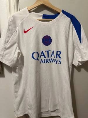 PSG training kit authentic  - Vit PSG matchtröja från Nike med blått Paris Saint-Germain-emblem på bröstet, röd Nike-logga och blå detalj på axeln. Tröjan har tryckt Qatar Airways-logga framtill och är tillverkad i ett lätt, ventilerande material perfekt för fotboll.