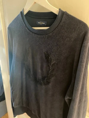 Svart sweatshirt från Fred Perry - Snygg svart sweatshirt från Fred Perry med broderad lagerkrans på bröstet. Tröjan har rund halsringning och ribbade muddar vid ärmslut och nederkant. Perfekt för en stilren och avslappnad look.