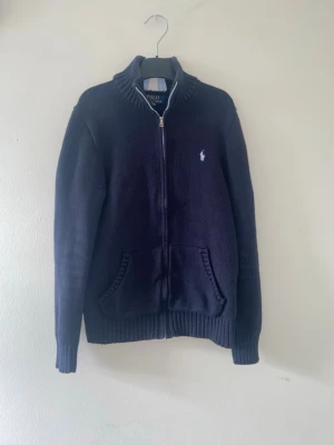 Mörkblå ziptröja från Polo Ralph Lauren - Mörkblå stickad ziptröja från Polo Ralph Lauren med vit broderad logga på bröstet. Tröjan har hög krage med randig detalj på baksidan, två fickor fram och ribbade muddar. Perfekt för lager-på-lager och chill stil. Pris kan diskuteras 