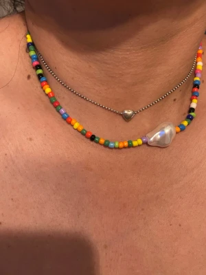 Färgglatt pärlhalsband med stor pärla - Ett halsband med små färgglada pärlor i regnbågens färger och en stor oregelbunden vit pärla som detalj. Halsbandet har en lekfull och trendig stil, perfekt för att ge din outfit en färgklick. Materialet är plastpärlor och en stor sötvattenspärla.