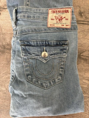 True Religion Joey Low Rise Flare Jeans - Säljer ett par ljusblå True Religion Joey jeans med låg midja och utsvängda ben. Klassisk bakficka med dekorativa sömmar samt silverfärgad knapp.  Som man ser på näst sista bilden är det en väldigt ljus fläck som knappt syns och som säkert går bort med lite fläckborttagning men även på sista bilden så är den lite söndriga då jag är kort och har gått på dem för att dem var för långa men ingenting som man riktigt tänker på när dem väl är på❤️