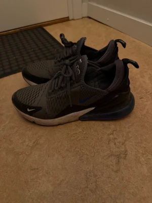Nike Air Max 270 svarta sneakers - Säljer ett par Nike Air Max 270 sneakers i svart och mörkgrått med blå detaljer. Skorna har en luftig mesh-överdel, synlig Air Max-dämpning i hälen och klassisk Nike-logga på sidan. Snygg och sportig design med snörning och låg profil.