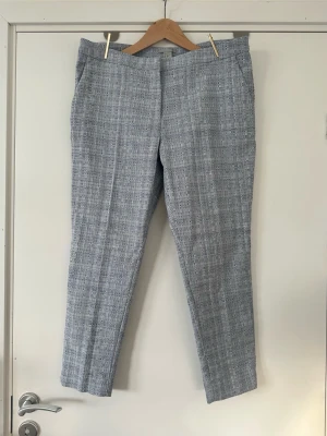 H&M - blå vita byxor - storlek 42 - Snygga vit-blå / ljusblå chinos / kostymbyxor / byxor från H&M i storlek 42. Vackert strukturvävt rutmönster i tyget och klassiska raka ben med lätt pressveck. Diskreta fickor i sidorna och passpoalfickor bak. Perfekta för en stilren och modern look. Mycket gott skick, som nya! 