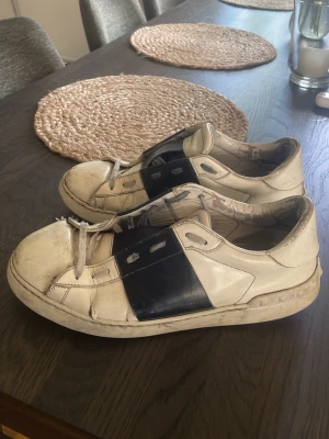 Valentino vita sneakers med svart detalj - Säljer ett par vita sneakers från Valentino med bred svart rem över mitten. Skorna har klassisk låg profil, rund tå och vita snören. Tillverkade i läder med diskret logga på plösen. Perfekta för dig som gillar stilrena men ändå unika sneakers.