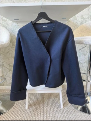 Mörkblå cardigan från Gina Tricot - Stilren mörkblå cardiganjacka från Gina Tricot med öppen front och dekorativa sömmar längs kanterna. Jackan har långa ärmar med breda uppvik och är gjord i ett mjukt, tjockare tyg som ger en snygg och avslappnad look.