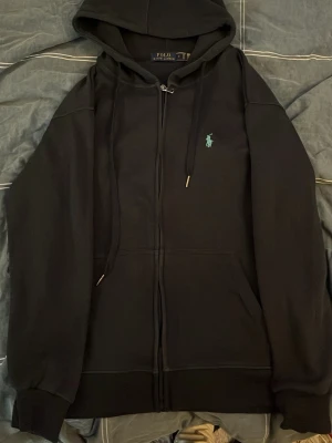 Marinblå hoodie från Polo Ralph Lauren - Säljer en marinblå hoodie från Polo Ralph Lauren med dragkedja framtill och klassisk huva med snören. På bröstet finns den ikoniska broderade loggan i ljusblått. Hoodien har ribbade muddar och är tillverkad i mjukt bomullsmaterial.