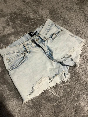 Ljusblå denimshorts från 157 - Säljer ett par ljusblå denimshorts från 157 med råa, fransiga kanter och slitna detaljer framtill. Shortsen har klassisk femficksdesign, bälteshällor och knappgylf. Perfekta för varma dagar och en avslappnad look.