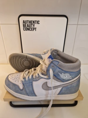 Nike Air Jordan 1 High blå/vit - Snygga Nike Air Jordan 1 High sneakers i blått och vitt med grå detaljer. Skorna har klassisk hög siluett, vit läderbas och blå mockadetaljer. Ikonisk Air Jordan-logga på sidan och vit sula. Perfekta för dig som vill sticka ut med stilrena och trendiga sneakers.