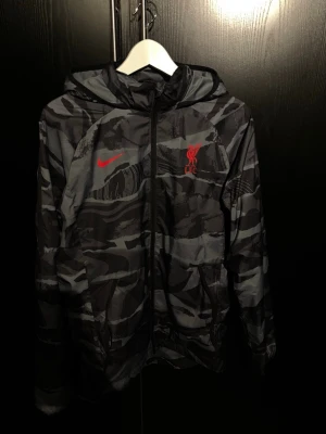 Nike x Liverpool camo vindjacka - Snygg vindjacka från Nike i samarbete med Liverpool FC. Jackan har ett grått och svart camomönster, röd Nike-logga och Liverpool FC-märke på bröstet. Utrustad med huva och dragkedja framtill. Perfekt för dig som vill sticka ut med sportig stil.