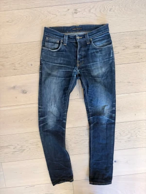 Blå slim fit jeans från Nudie Jeans - Snygga blå slim fit jeans från Nudie Jeans med slitna detaljer och klassisk femficksdesign. Jeansen har tydliga whiskers och kontrastsömmar, tillverkade i denim med en cool tvättad look. Perfekta för en avslappnad och trendig stil.