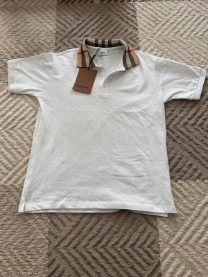 Vit pikétröja med rutig krage Burberry - Vit pikétröja från Burberry med klassisk rutig krage i beige, svart och rött. Tröjan har korta ärmar, knappar framtill och är tillverkad i bomull. Perfekt för en stilren och trendig look.