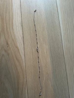 Zadig&voltaire necklace - Ett tunt armband i silverfärg med en unik mittdel formad som två vingar täckta av små glittrande stenar. Kedjan har små dekorativa pärldetaljer längs länken. Perfekt för dig som gillar stilrena och eleganta accessoarer.