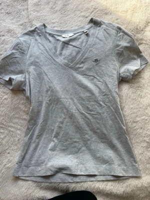 Grå v-ringad t-shirt från GANT - Snygg grå t-shirt från GANT i mjuk bomull. T-shirten har v-ringning, korta ärmar och en liten broderad logga på bröstet. Perfekt basplagg med en clean och klassisk look.