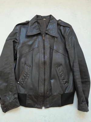 Svart vintage skinnjacka med dragkedja - Snygg vintage svart skinnjacka med klassisk krage, axelklaffar och ribbade muddar. Jackan har två snedställda fickor framtill och stängs med dragkedja. Snygga sömdetaljer både fram och bak ger en cool look.