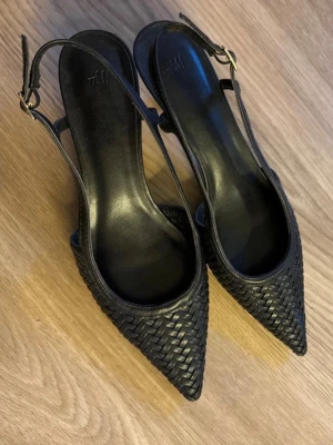 Svarta flätade pumps från H&M - Säljer ett par snygga svarta klackar från H&M med spetsig tå och flätad design. Skorna har en öppen häl med justerbar rem, aldrig använda pga för stora