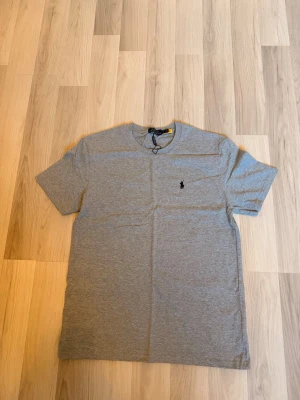 Grå t-shirt från Polo Ralph Lauren - Snygg grå t-shirt från Polo Ralph Lauren med klassisk rund hals och den ikoniska broderade loggan på bröstet. Tillverkad i mjuk bomull och har en enkel, clean look som passar till allt. Perfekt basplagg för en stilren vardagsoutfit.