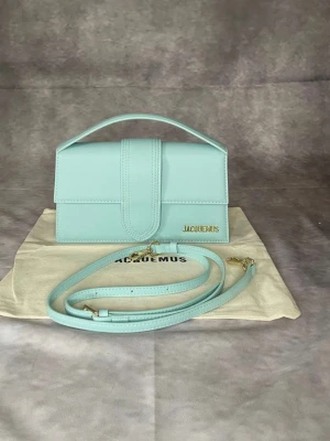 Ljusblå Jacquemus handväska - Superfin ljusblå handväska från Jacquemus i stilren, rektangulär modell. Väskan är i slätt läder med guldfärgad logga och har både handtag och avtagbar axelrem. Perfekt för dig som gillar trendiga accessoarer och vill sticka ut med en poppig färg.