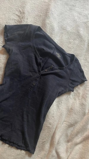 Svart ribbad topp från Hollister - Snygg svart ribbad topp från Hollister med korta ärmar och v-ringning. Toppen har knappar framtill och är i ett mjukt, stretchigt material som sitter tight. Perfekt till jeans eller kjol för en enkel men stilren look.