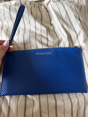 Blå kuvertväska Michael Kors - Snygg blå kuvertväska från Michael Kors i perforerat läder med silverfärgad dragkedja och handledsrem. Perfekt att bära till mobilen och småsaker. Diskret logga framtill och ljus textilinsida.