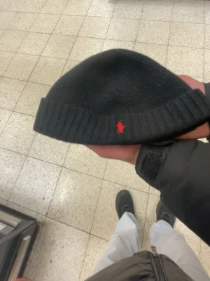 Svart mössa från Polo Ralph Lauren - Snygg svart mössa från Polo Ralph Lauren med klassisk röd logga broderad framtill. Mössan har ribbad kant och är tillverkad i mjukt material som sitter skönt på huvudet. Perfekt accessoar för kalla dagar.